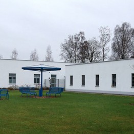 Zwei weiße, einstöckige Gebäude mit vergitterten Fenstern. Im Vordergund steht eine blaue Sitzgruppe auf grünem Rasen.