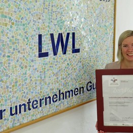 Eine junge Frau hält lächelnd ein gerahmtes Zertifikat in den Händen.