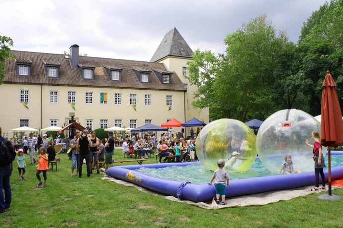 Ein Wasserfläche mit spielenden Kindern in transparenten Riesenwasserbällen vor einem schlossähnlichen Gebäude.