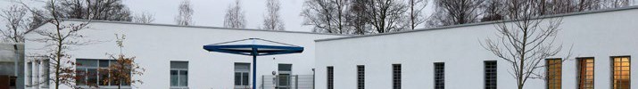 Zwei weiße, einstöckige Gebäude mit vergitterten Fenstern und zwei blaue Sitzgruppen auf grünem Rasen. Die Bäume im Hintergrund tragen kein Laub.