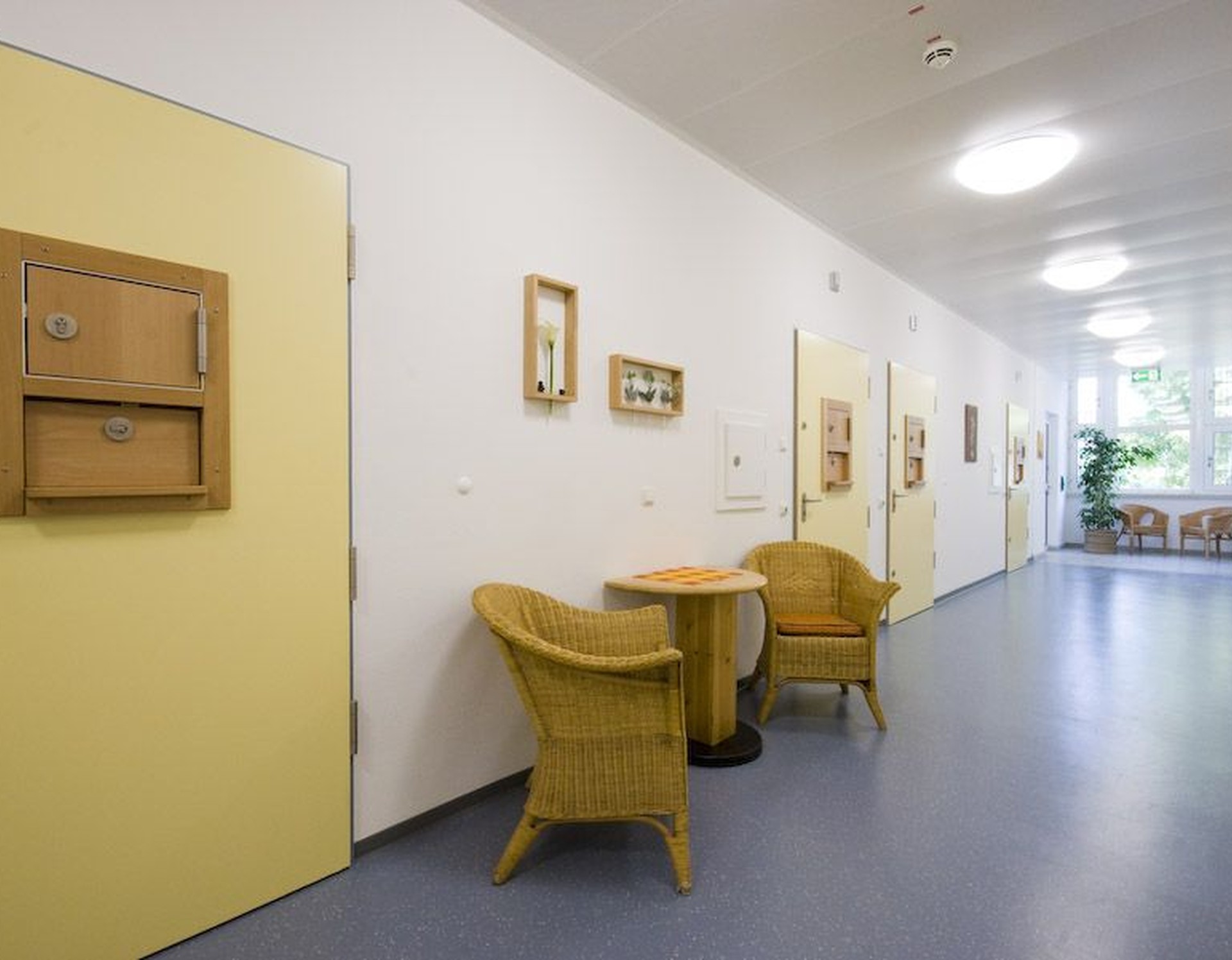 Ein heller Flur einer geschlossenen Therapiestation mit mehreren geschlossenen gelben Türen und mehreren gemütlichen Korbstühlen.